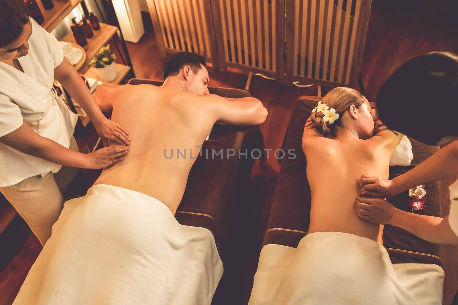 Caucasian couple customer enjoying relaxing anti-stress spa massage and pampering with beauty skin recreation leisure in warm candle lighting ambient salon spa at luxury resort or hotel. Quiescent