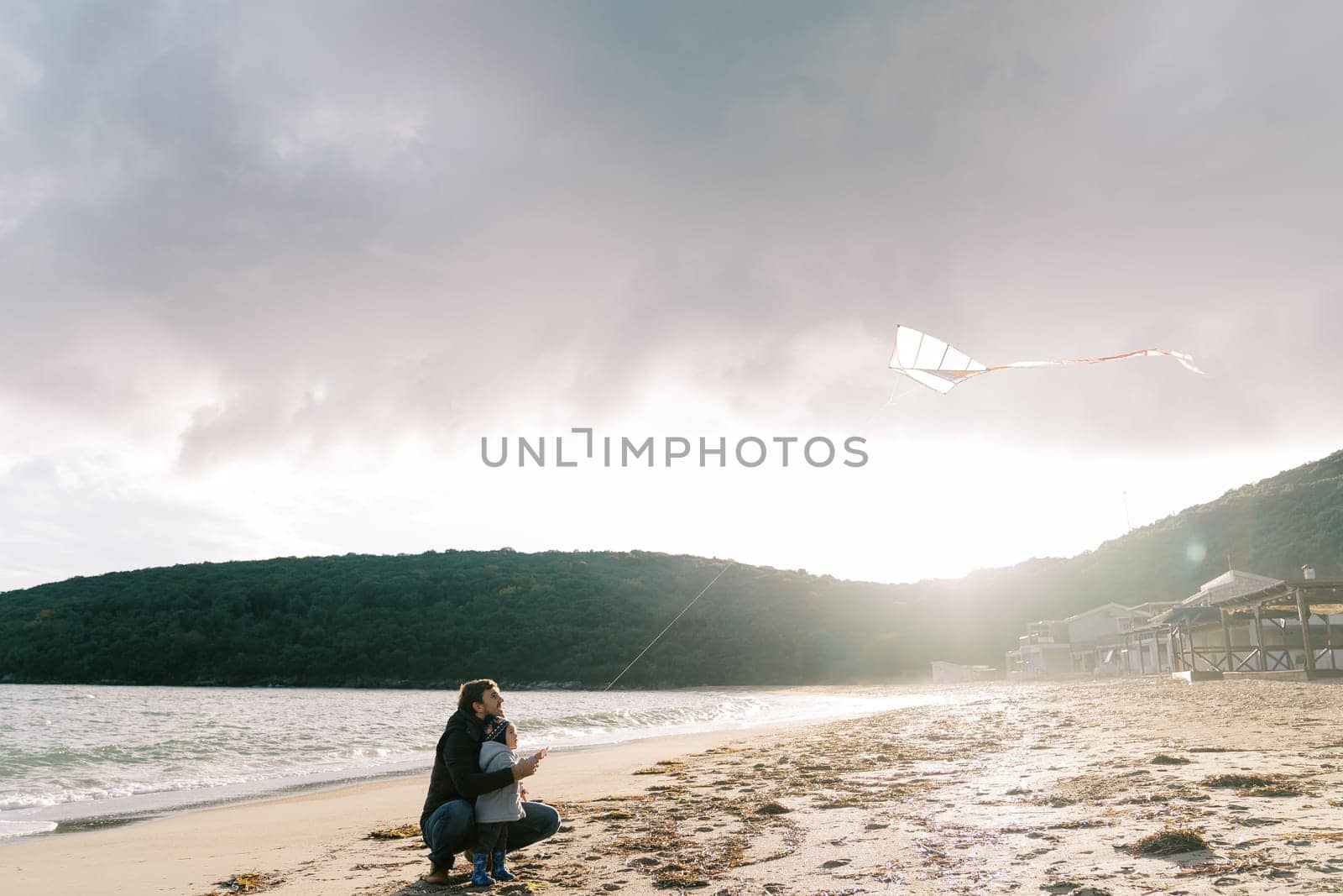 Dad with a little girl flying a kite sitting on the beach by the sea. High quality photo