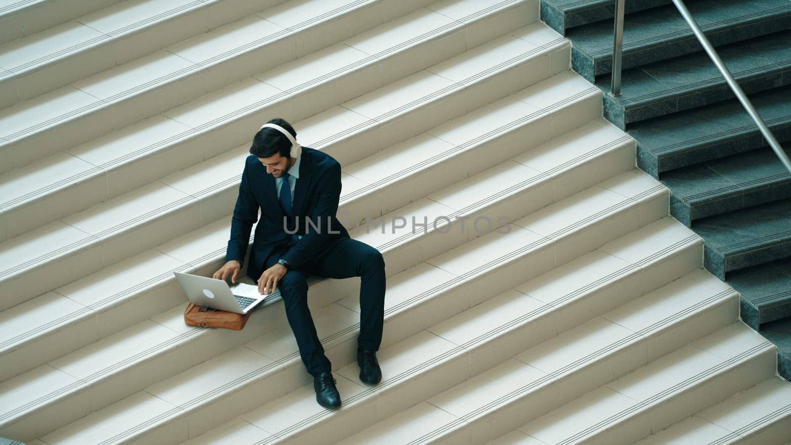 Relaxing smart business man listening music from headphone while sitting at stair. Happy manager smiling while dancing or moving to music. Investor receive good news, getting promotion. Exultant.