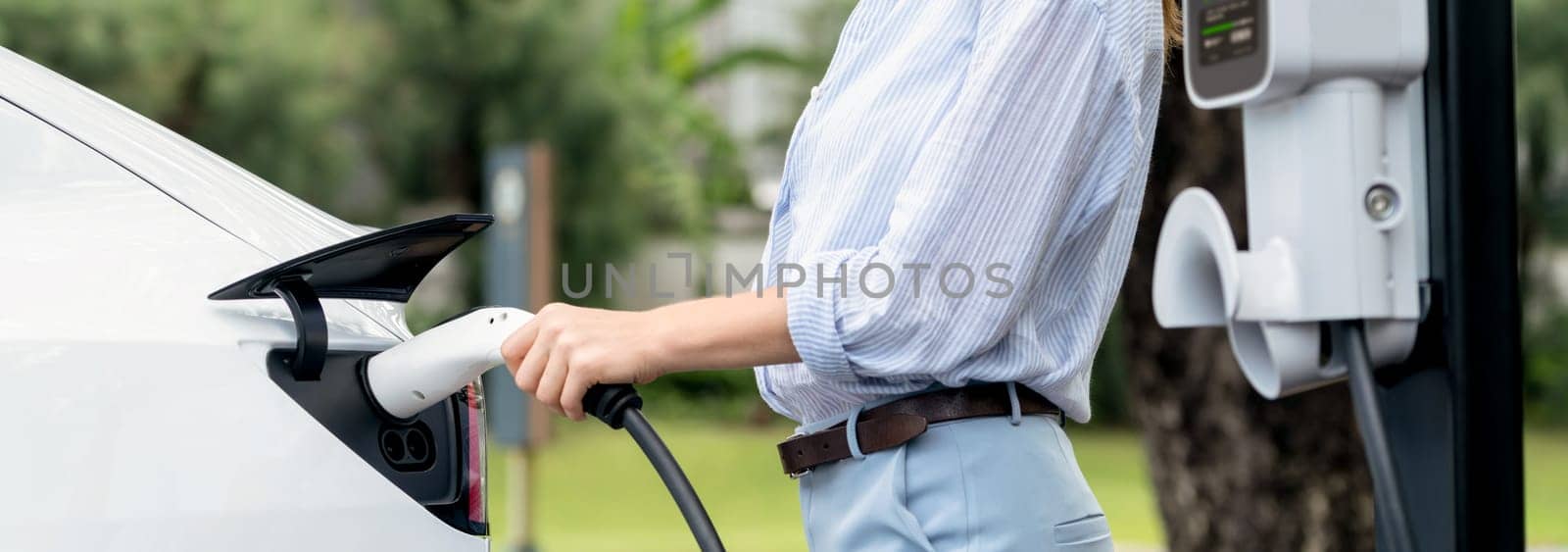 Young woman recharge EV electric vehicle's battery from EV charging station in outdoor green city park scenic. Eco friendly urban transport and commute with eco friendly EV car travel. Panorama Exalt