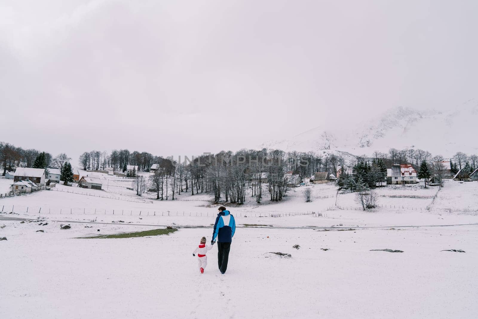 Father and a small child walk holding hands through a snowy pasture towards the village. Back view by Nadtochiy