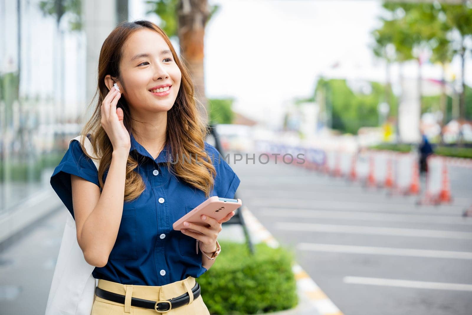 Happy young woman multitasking with wireless earbuds and smartphone in an urban environment. She's a portrait of a modern entrepreneur.