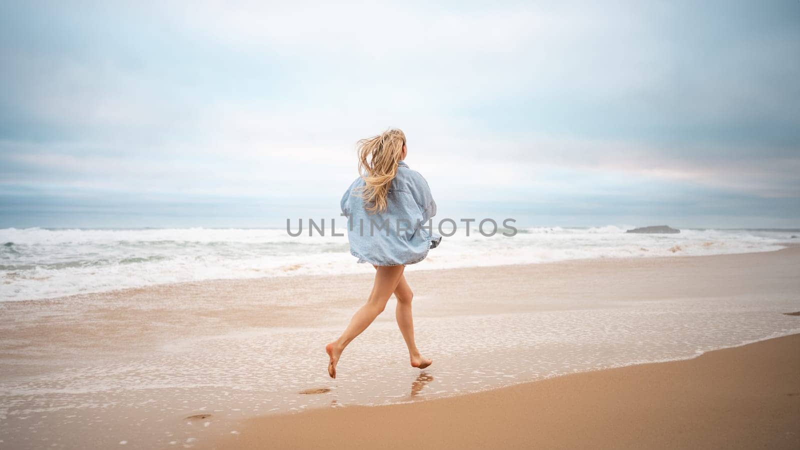 Full length of carefree woman with blond hair running along sea on sandy beach. Rear view of female tourist in denim shirt on summer vacation. Excited lady is on beach holiday.