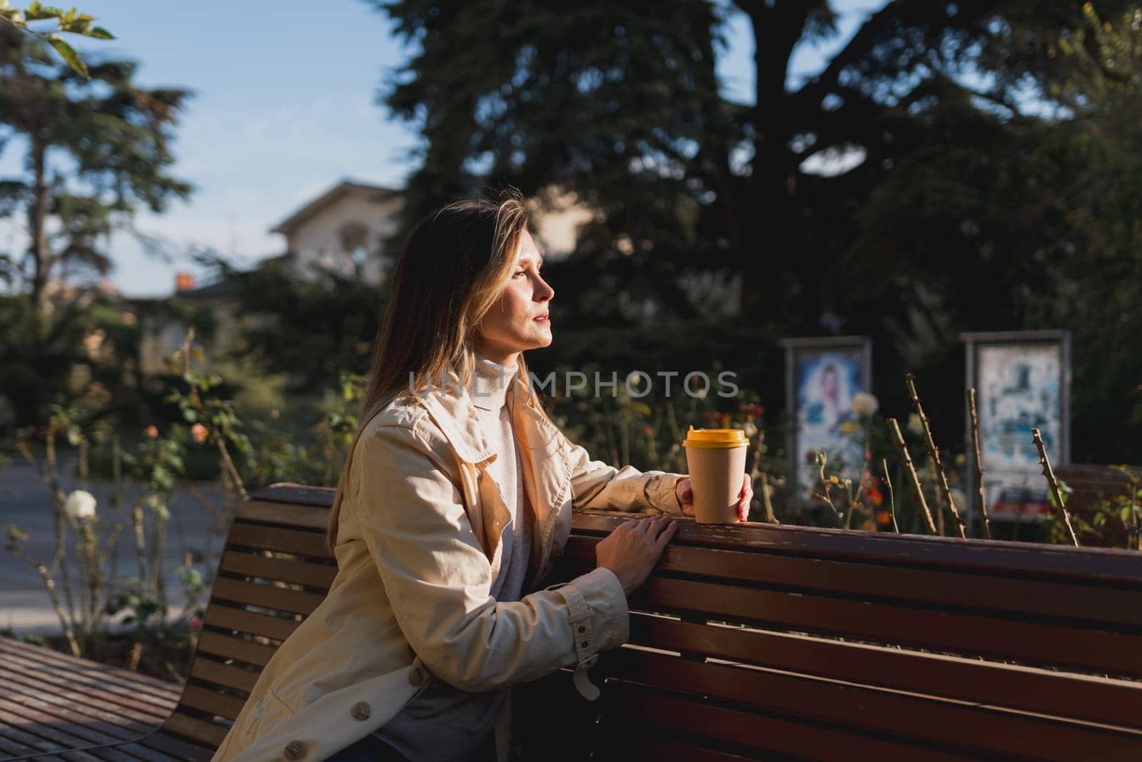 Woman drinks from cup on wooden bench. She is wearing a white shirt enjoying her beverage. The bench is located in a park setting, with trees in the background. by Matiunina