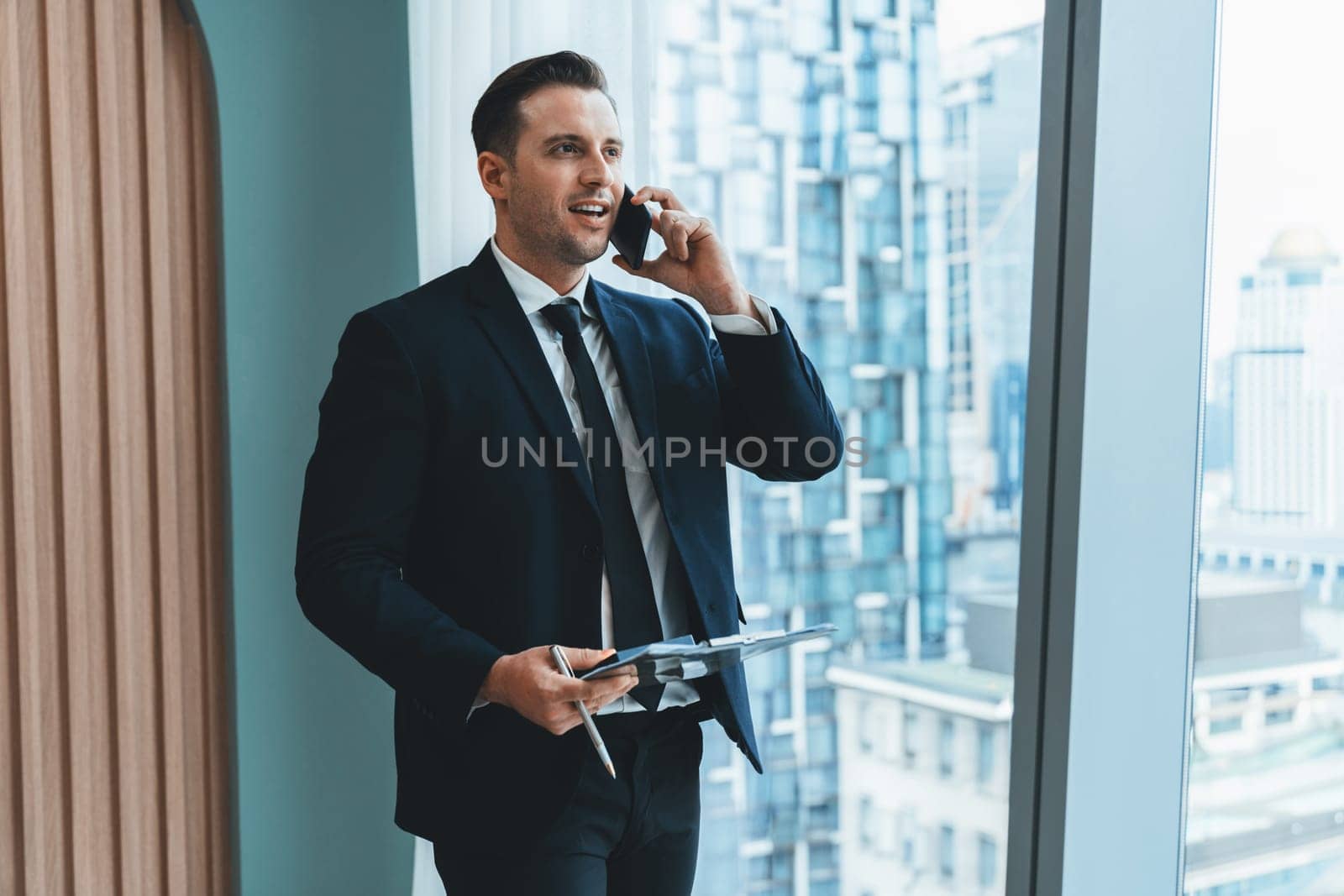 Portrait of professional businessman stand near window with skyscraper while phone calling and holding laptop. Busy executive manager holding laptop while multitasking working. Closeup. Ornamented.