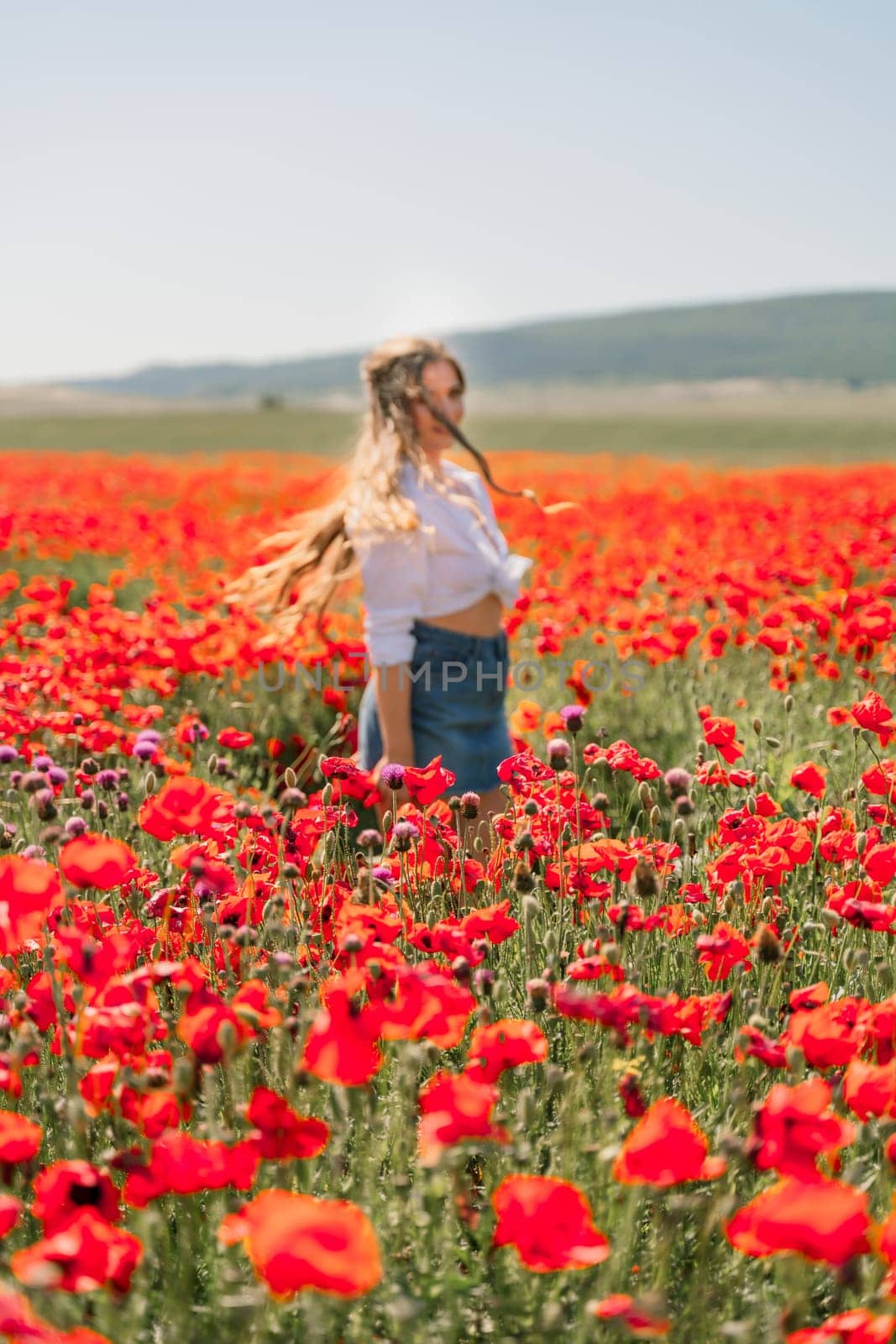 Happy woman in a poppy field in a white shirt and denim skirt with a wreath of poppies on her head posing and enjoying the poppy field