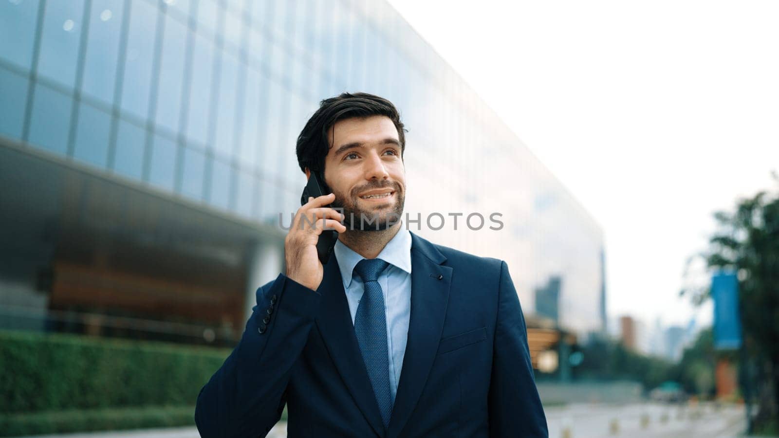 Skilled caucasian project manager calling phone while walking at city with blurred background. Skilled businessman using smart phone talking to project manager with blurred background. Exultant.