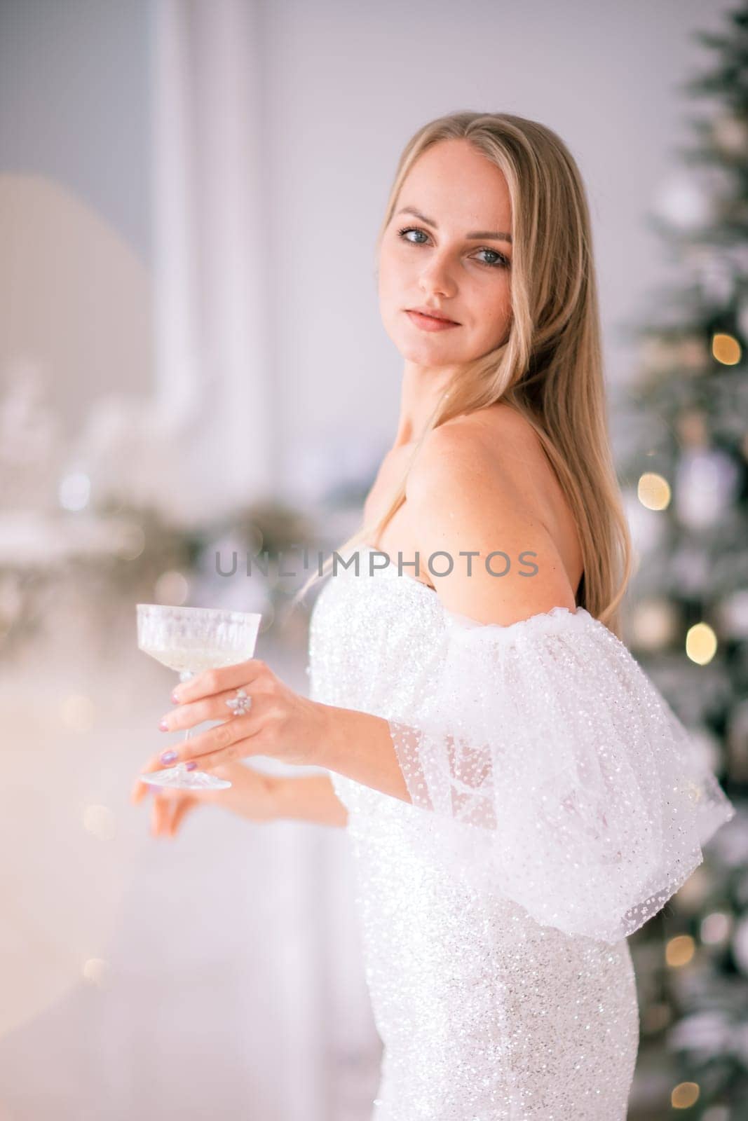 The blonde in the Christmas room. A beautiful blonde woman in a shiny light short dress with a train stands in a beautiful bright room decorated with a festive interior with a Christmas tree