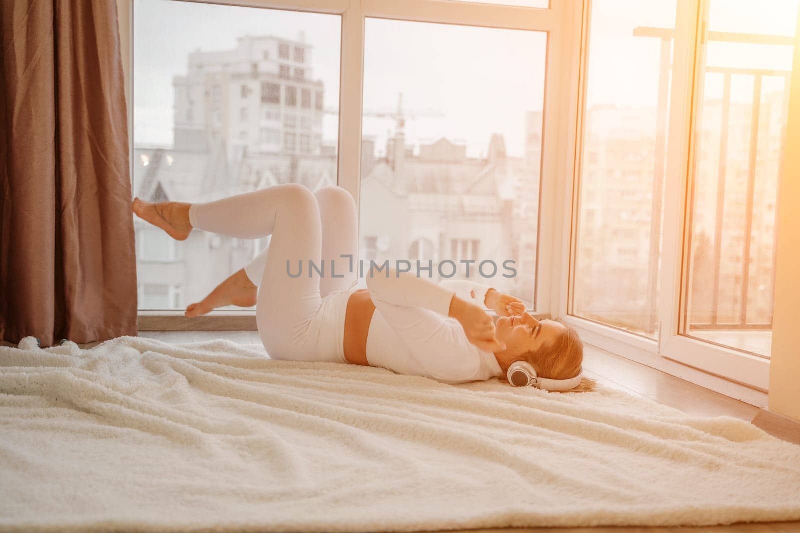 Side view portrait of relaxed woman listening to music with headphones lying on carpet at home. She is dressed in a white tracksuit