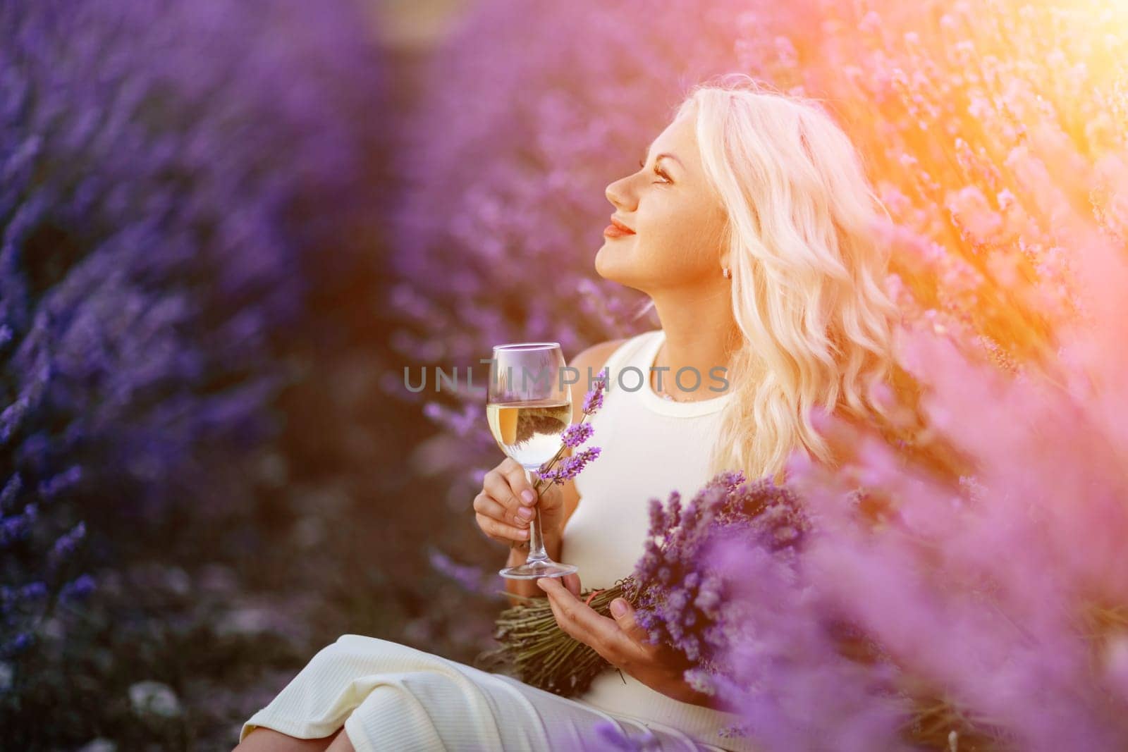Blonde lavender field holds a glass of white wine in her hands. Happy woman in white dress enjoys lavender field picnic holding a large bouquet of lavender in her hands . Illustrating woman's picnic in a lavender field. by Matiunina