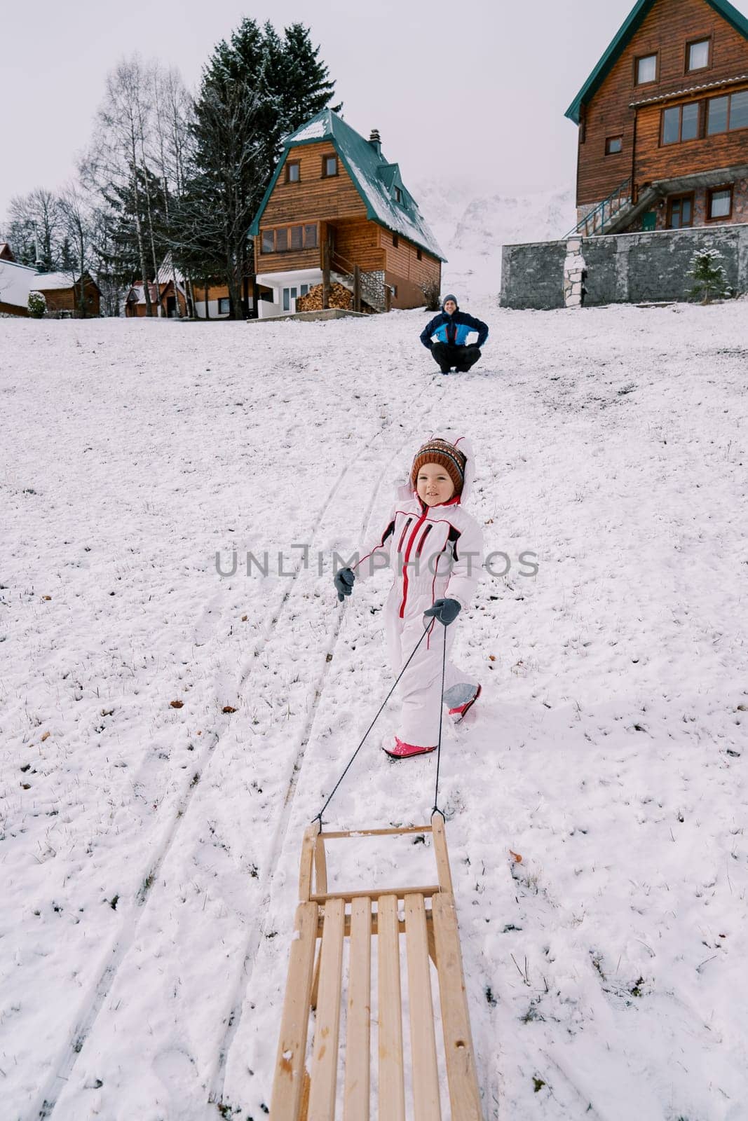 Little girl climbs a hill with her dad sitting on it with a sled on a rope, looking back. Back view. High quality photo