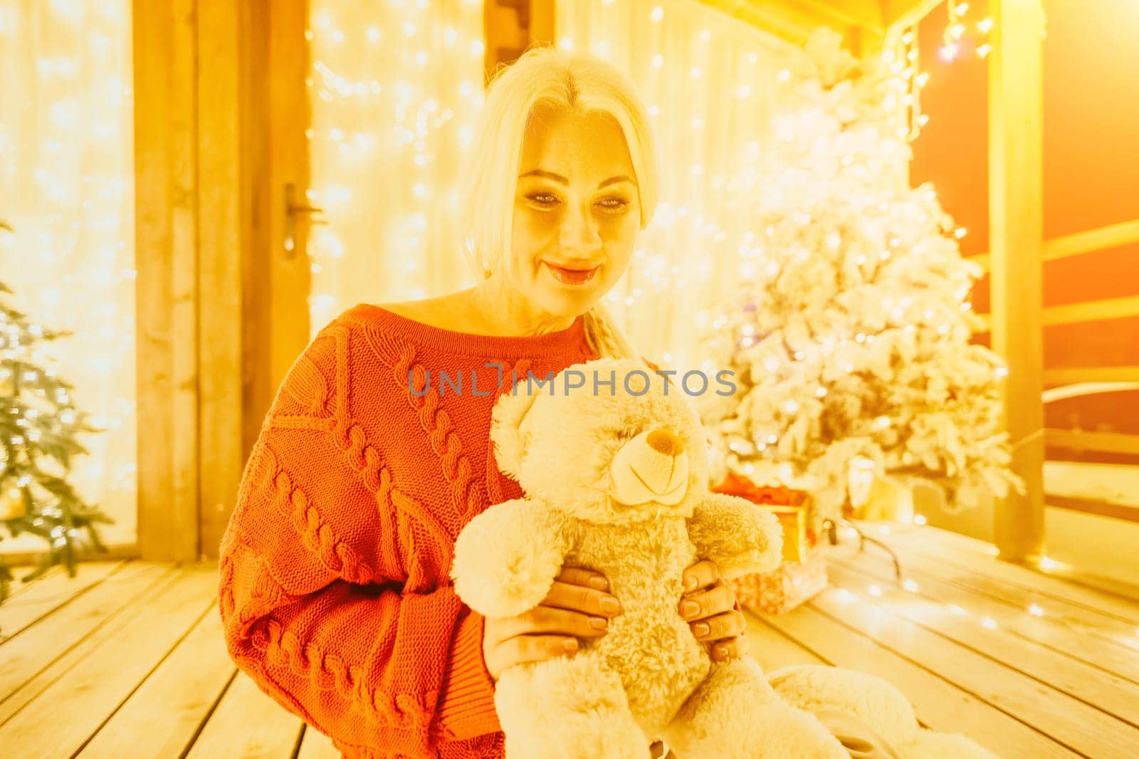 Happy smiling Woman in a red sweater sits on a festive porch, holding a teddy bear next to Snow-covered Christmas tree with bright white lights, exuding contented enjoyment in a cozy, cheerful smile by panophotograph