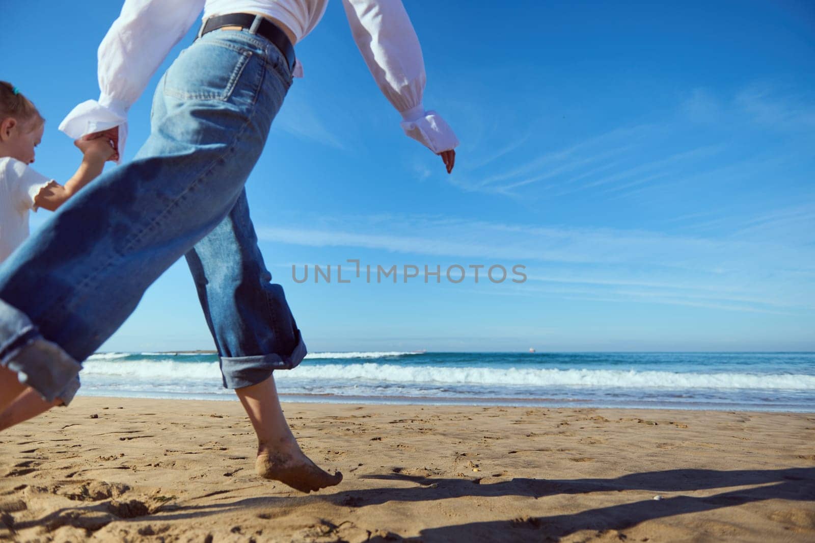 Close-up portrait of a Caucasian young mother and her little child girl holding hands while walking running barefoot on the sandy beach, dressed together in white shirt and blue denim jeans