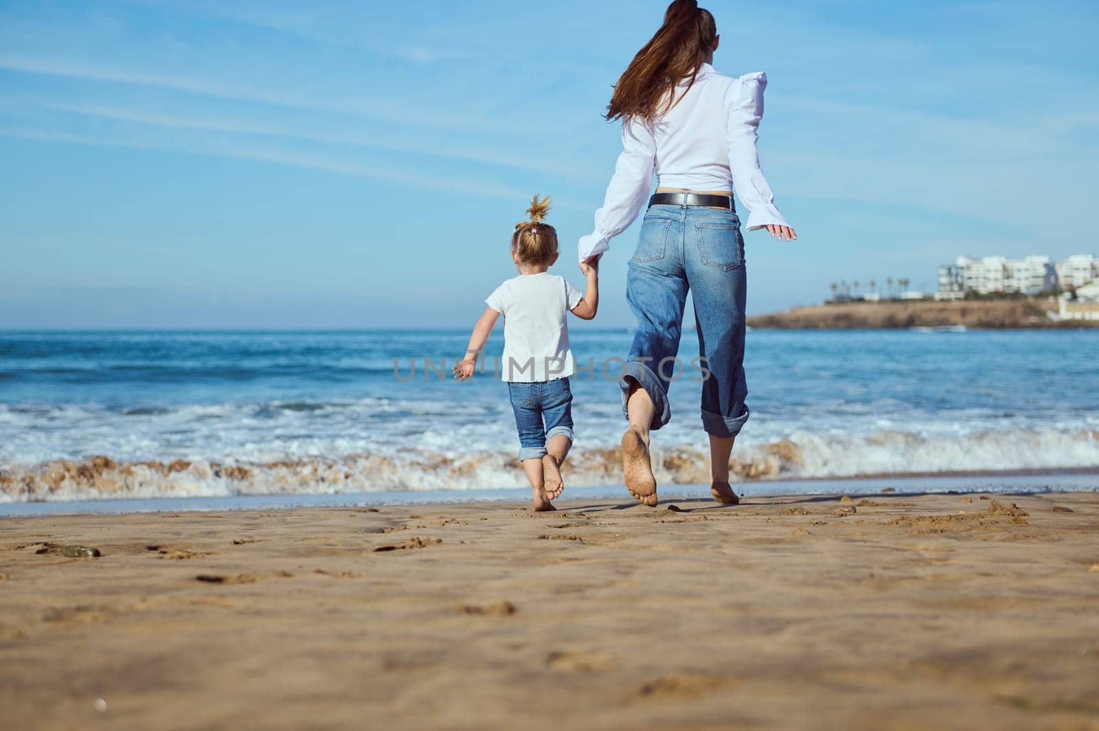 Full length rear portrait of a Caucasian young mother and her little child girl holding hands while walking running barefoot on the sandy beach, dressed together in white shirt and blue denim jeans