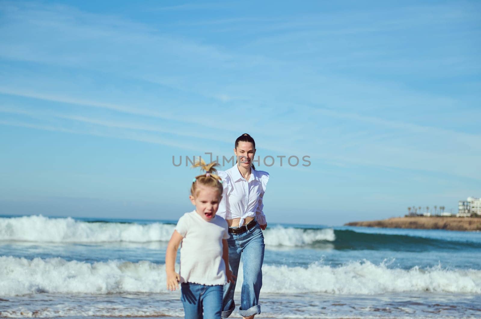 Young mother and little daughter playing together, running along a tropical beach, playing with waves crashing on the shore, enjoying happy time together. Family relationships. Carefree childhood