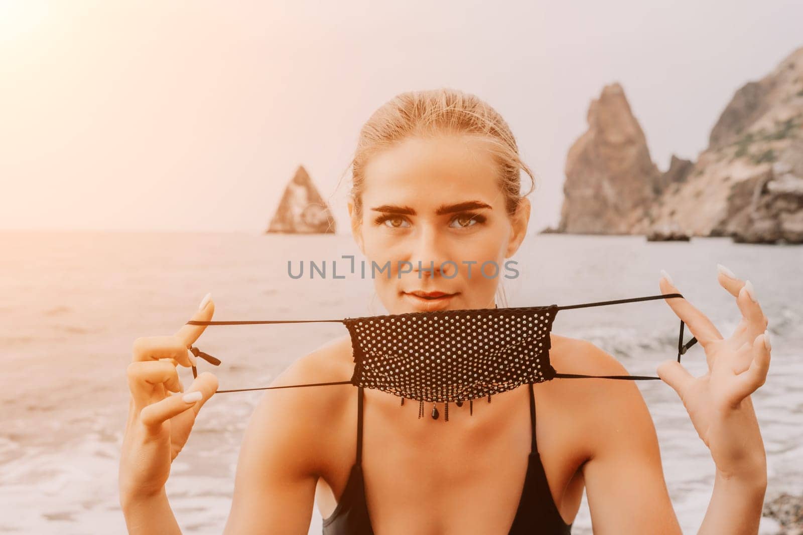 Woman travel portrait. Happy woman with long hair poses on a red volcanic rock at the beach. Close up portrait cute woman in black bikini, smiles at the camera, with the sea in the background. by panophotograph