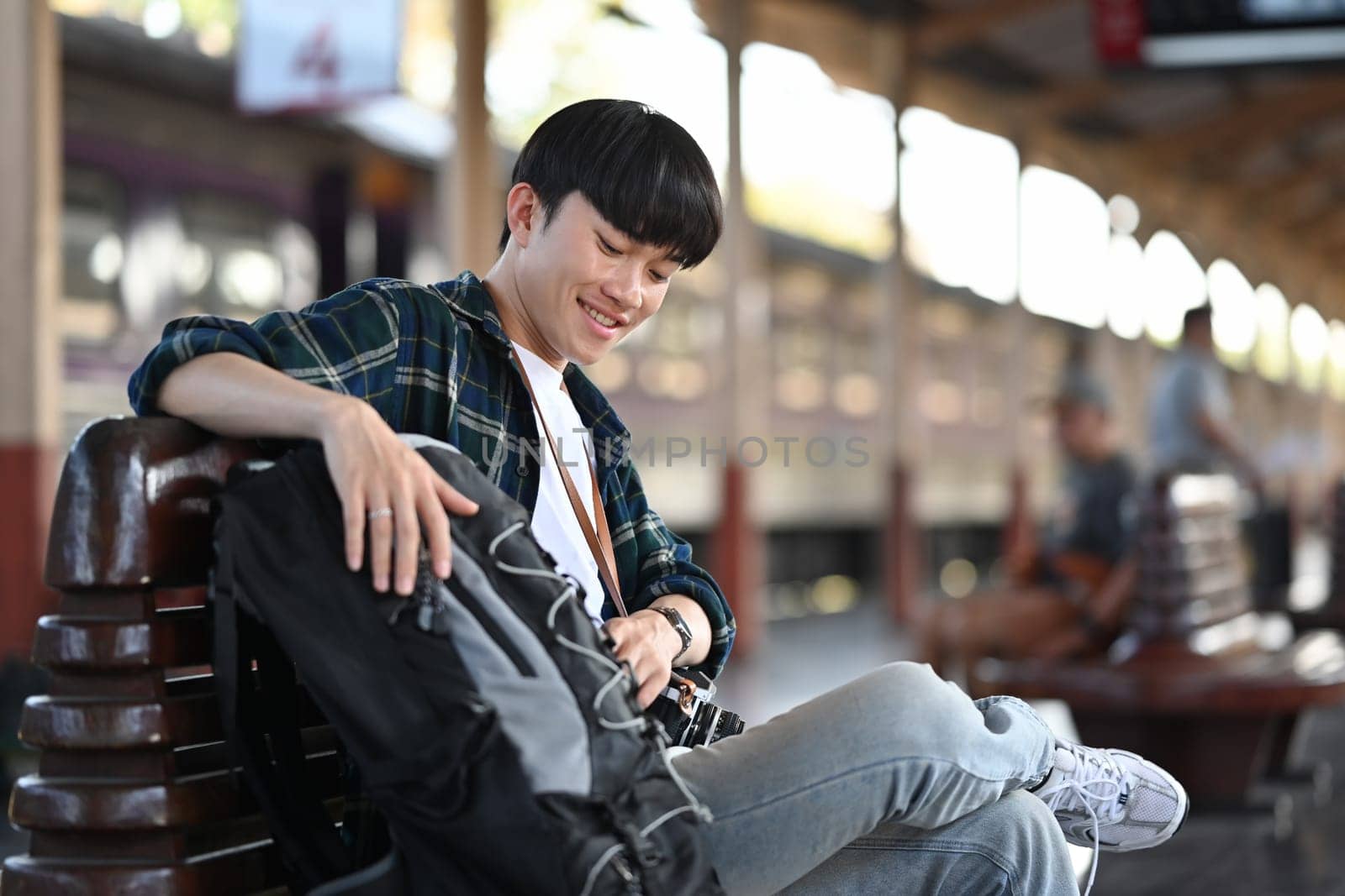 Young man backpacker sitting on a bench waiting train at railway platform. Travel and vacations concept.