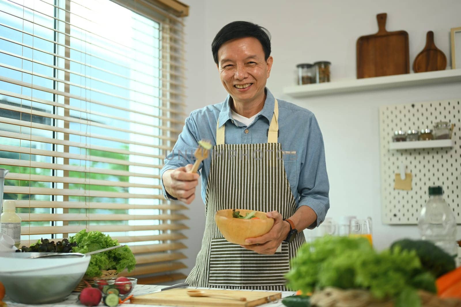 Positive middle age man in apron holding a bowl of salad and smiling at camera. Healthy eating concept.