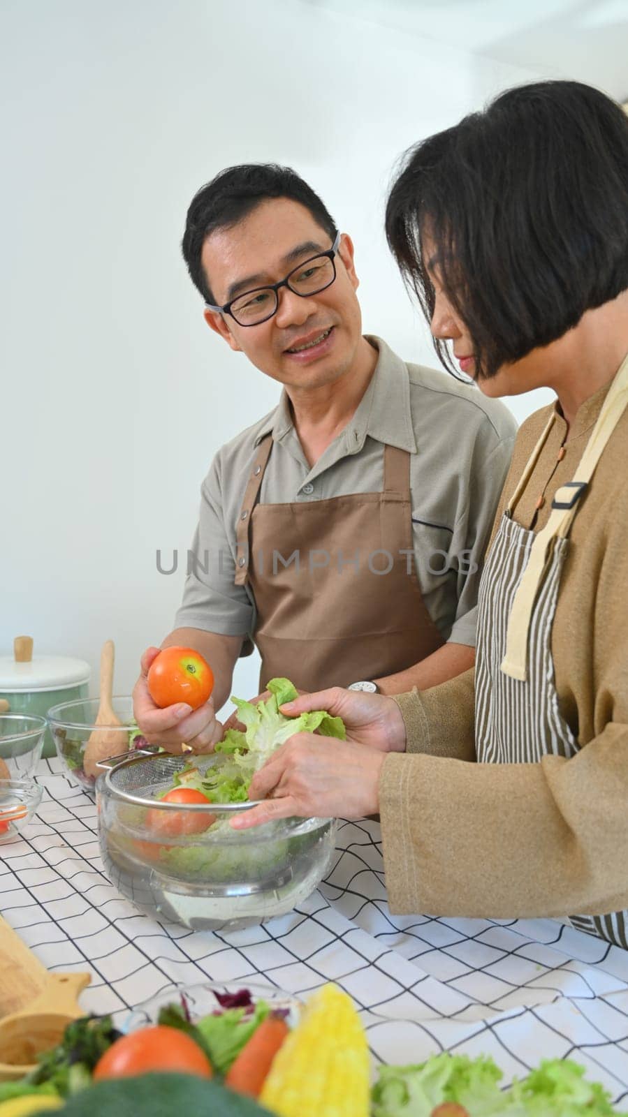Happy senior couple having conversation while cooking healthy vegan meal together in kitchen by prathanchorruangsak