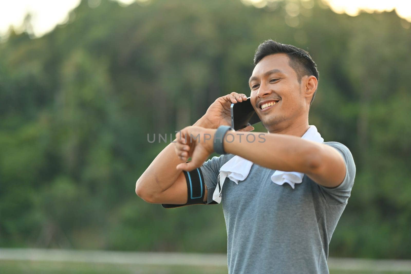 Smiling millennial man in sports clothes talking on mobile phone and checking time on watch by prathanchorruangsak