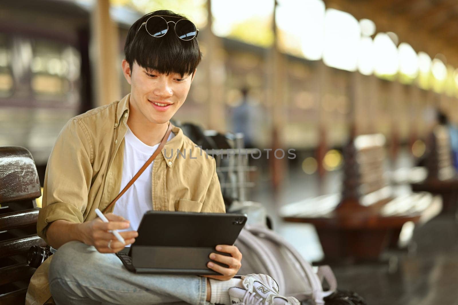 Male traveler using digital tablet while sitting on a bench at the train station by prathanchorruangsak