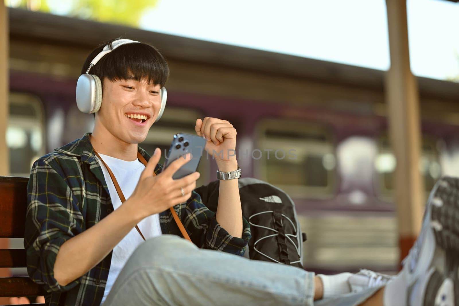 Cheerful asian man in headphone sitting on bench and using mobile phone while waiting for train.