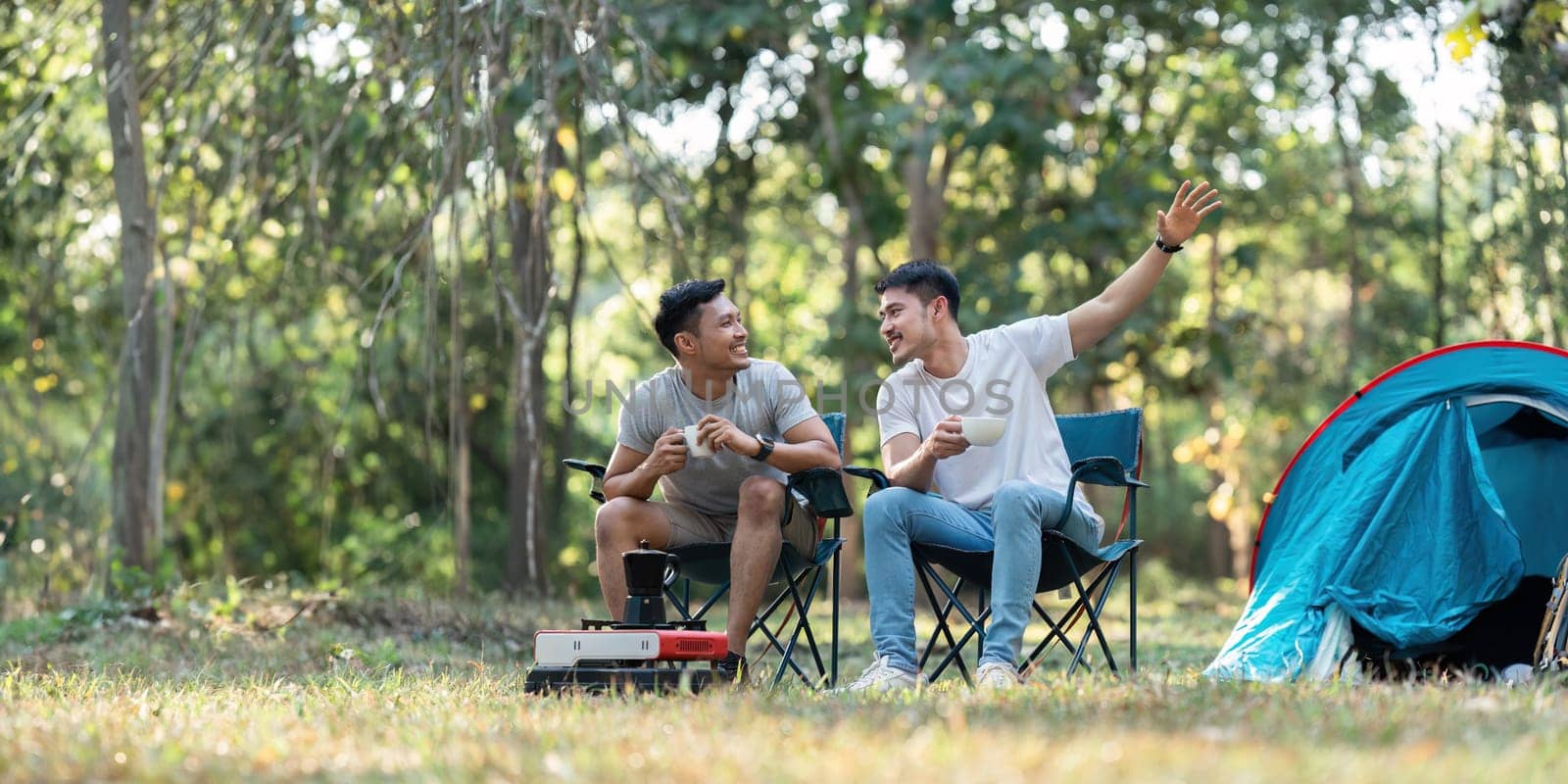 Gay LGBTQIA couple sitting on picnic chair drinking tea and coffee while camping on vacation holiday.