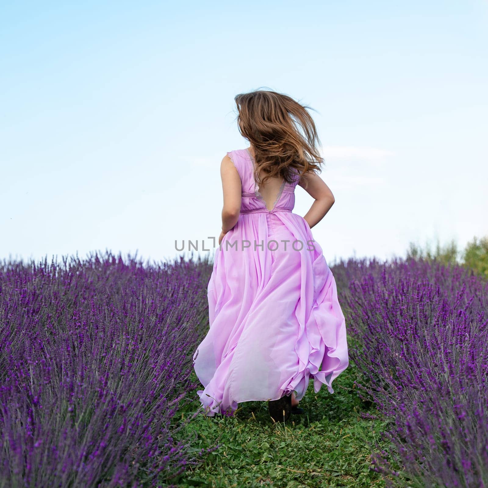 A young girl in a purple long dress runs between the lavender rows on the grass. by Niko_Cingaryuk