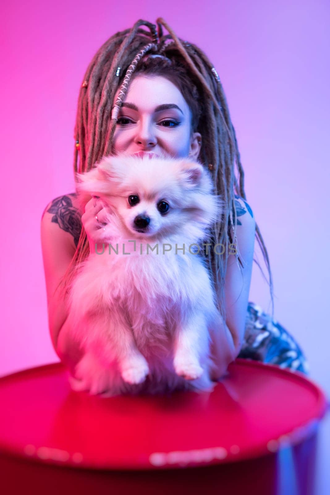 cool girl with braided dreadlocks on her head with a cute Pomeranian dog in neon light on a light background