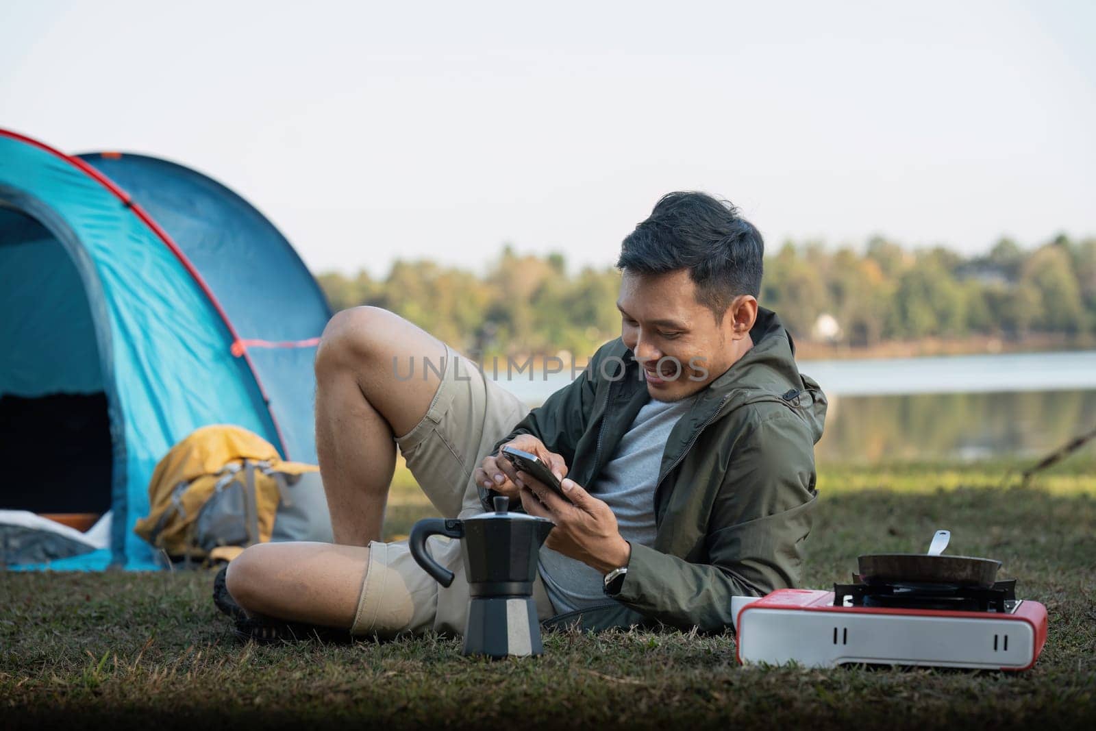 Young smiling man sitting near tent with smartphone and working remotely while camping in nature during adventure trip in forest by nateemee