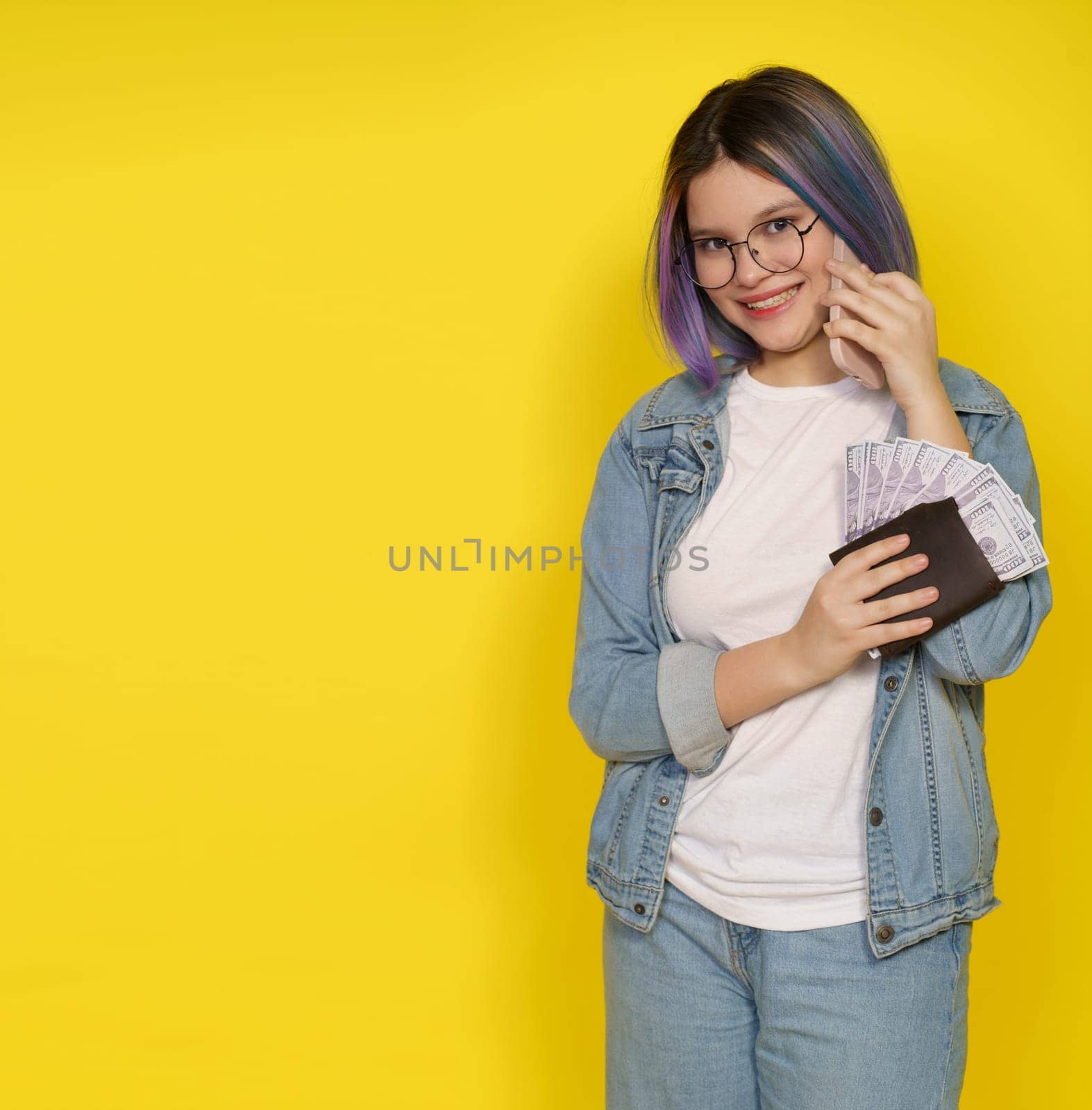 Financial Independence Associated With Managing Money In Modern And Confident Manner. Happy Smiling Teenager Girl With Wallet Full Of Money And Mobile Phone Isolated On Yellow With Copy Space. High quality photo