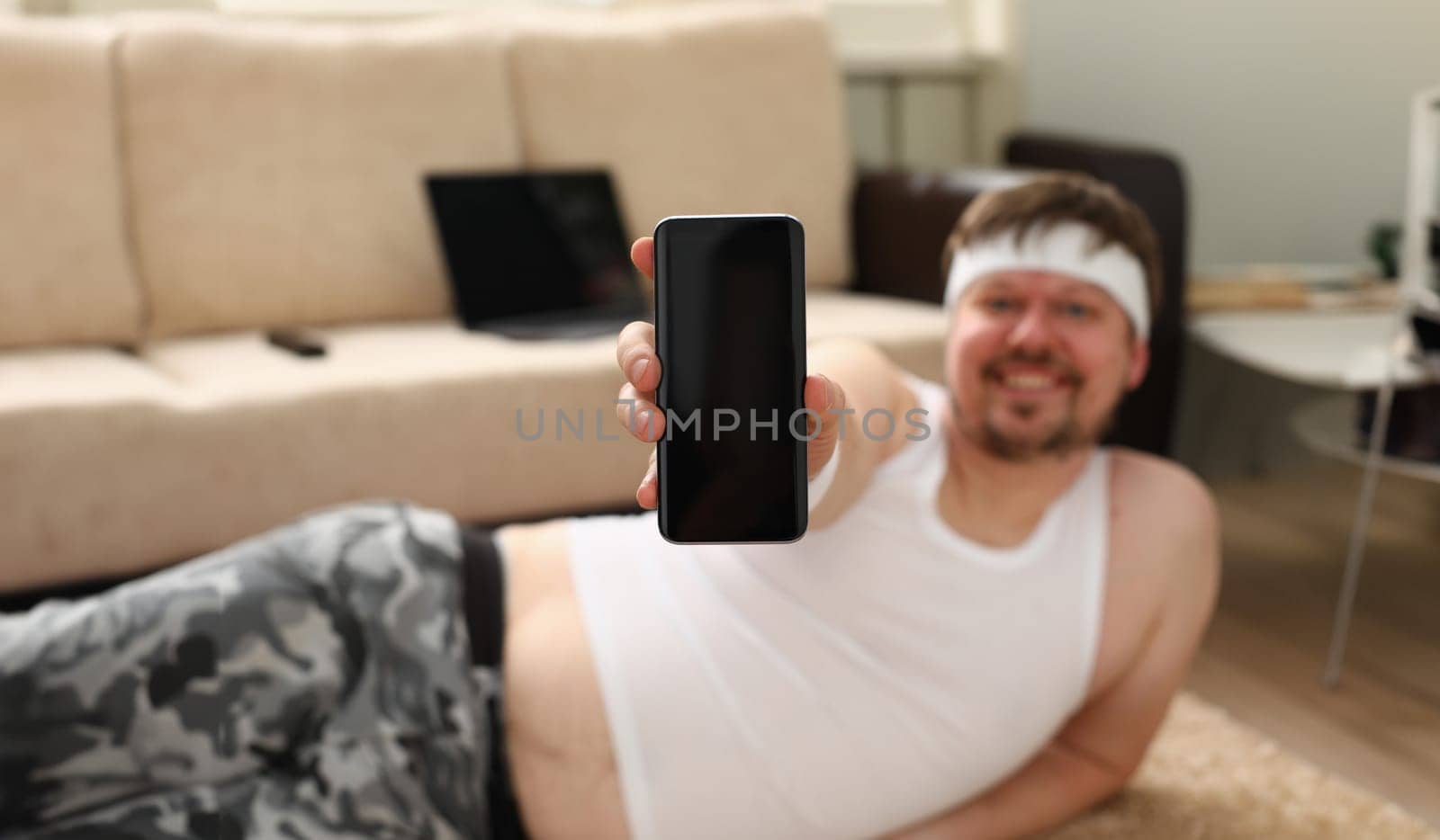 A young cute fat man with overweight does the first fitness workout at home demonstrates a smartphone in the camera with a training program holds in his hand.