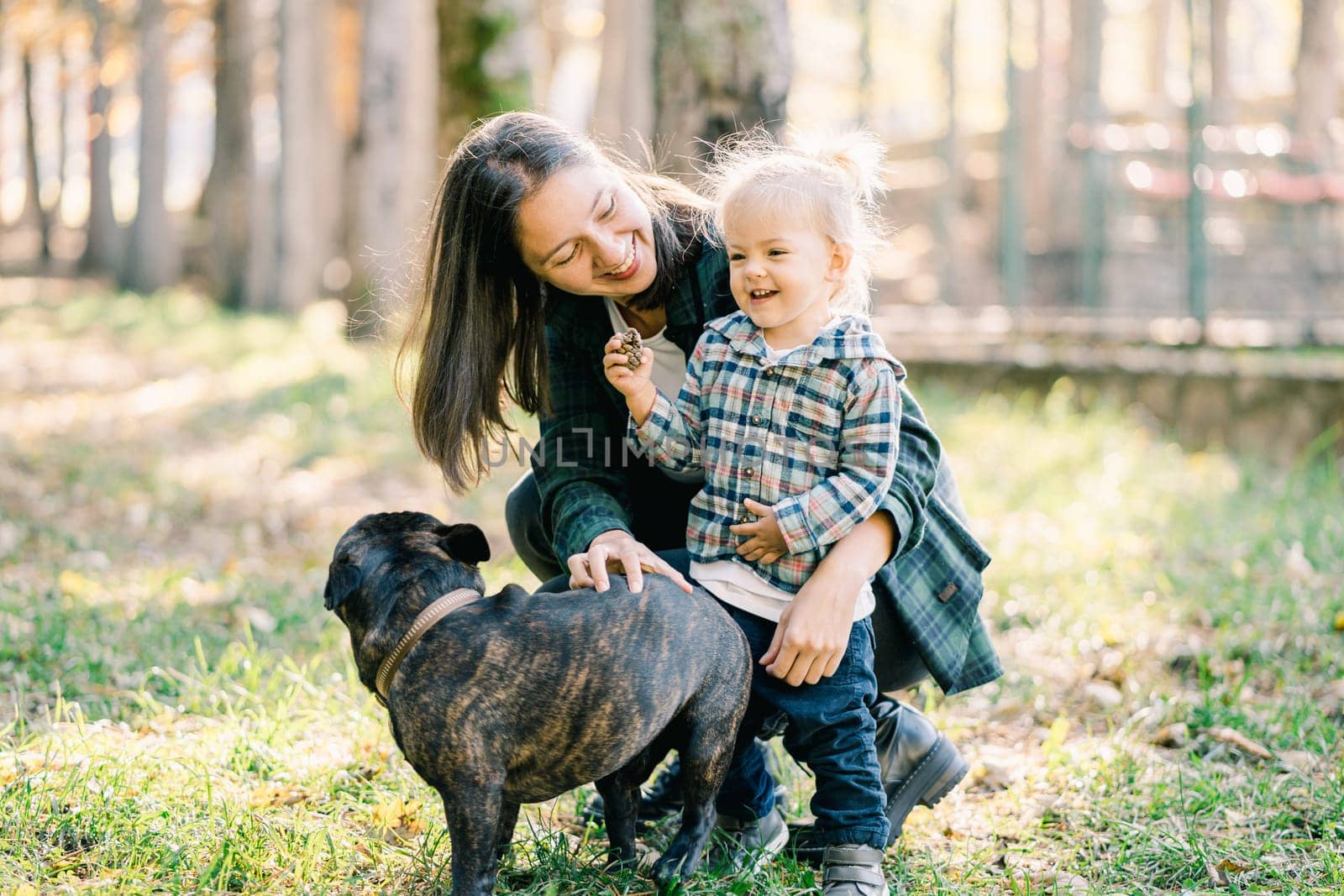 Smiling mother hugging little girl squatting next to her and petting dog in park. High quality photo