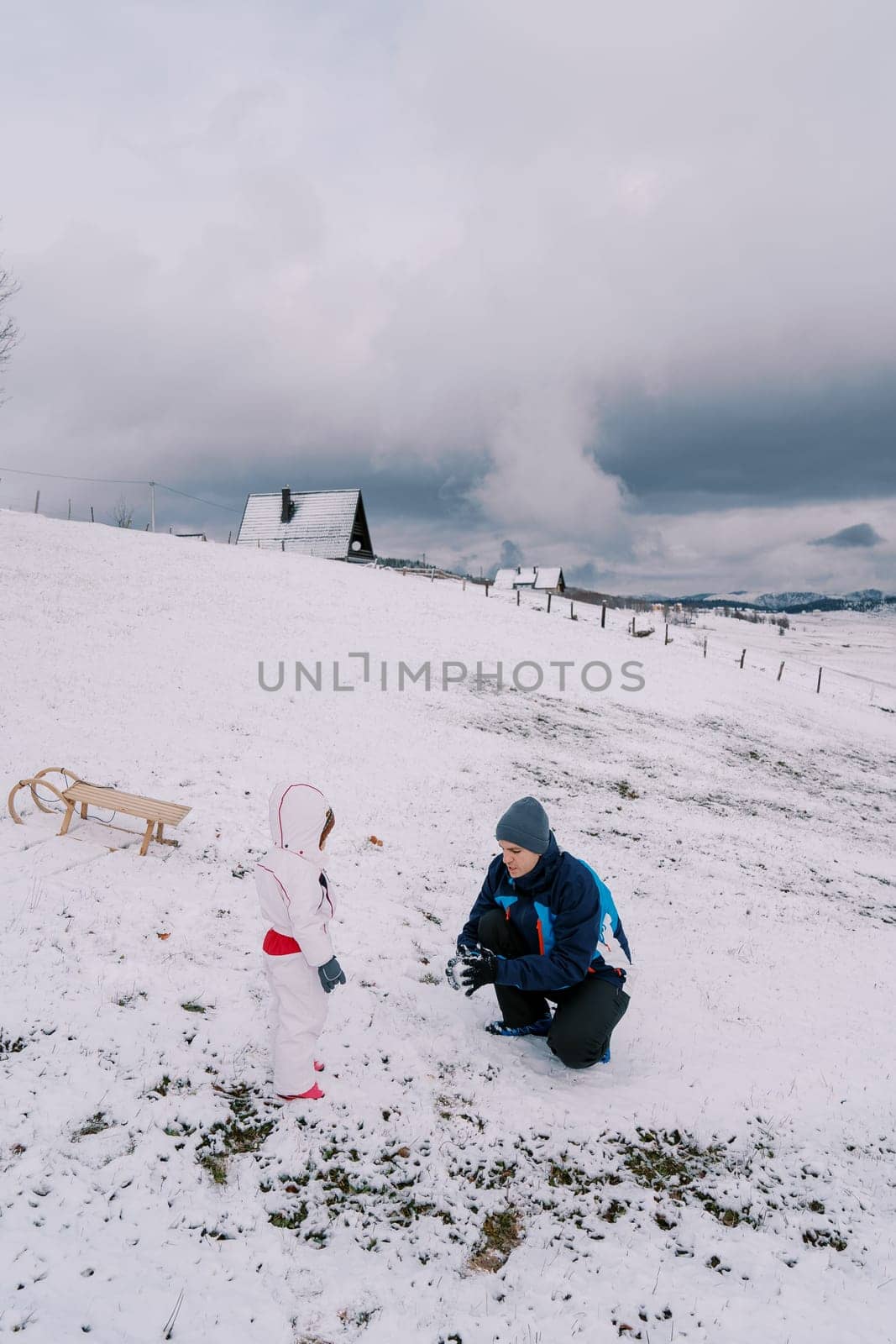 Small child looks at his dad squatting and making a snowball on a hill. High quality photo