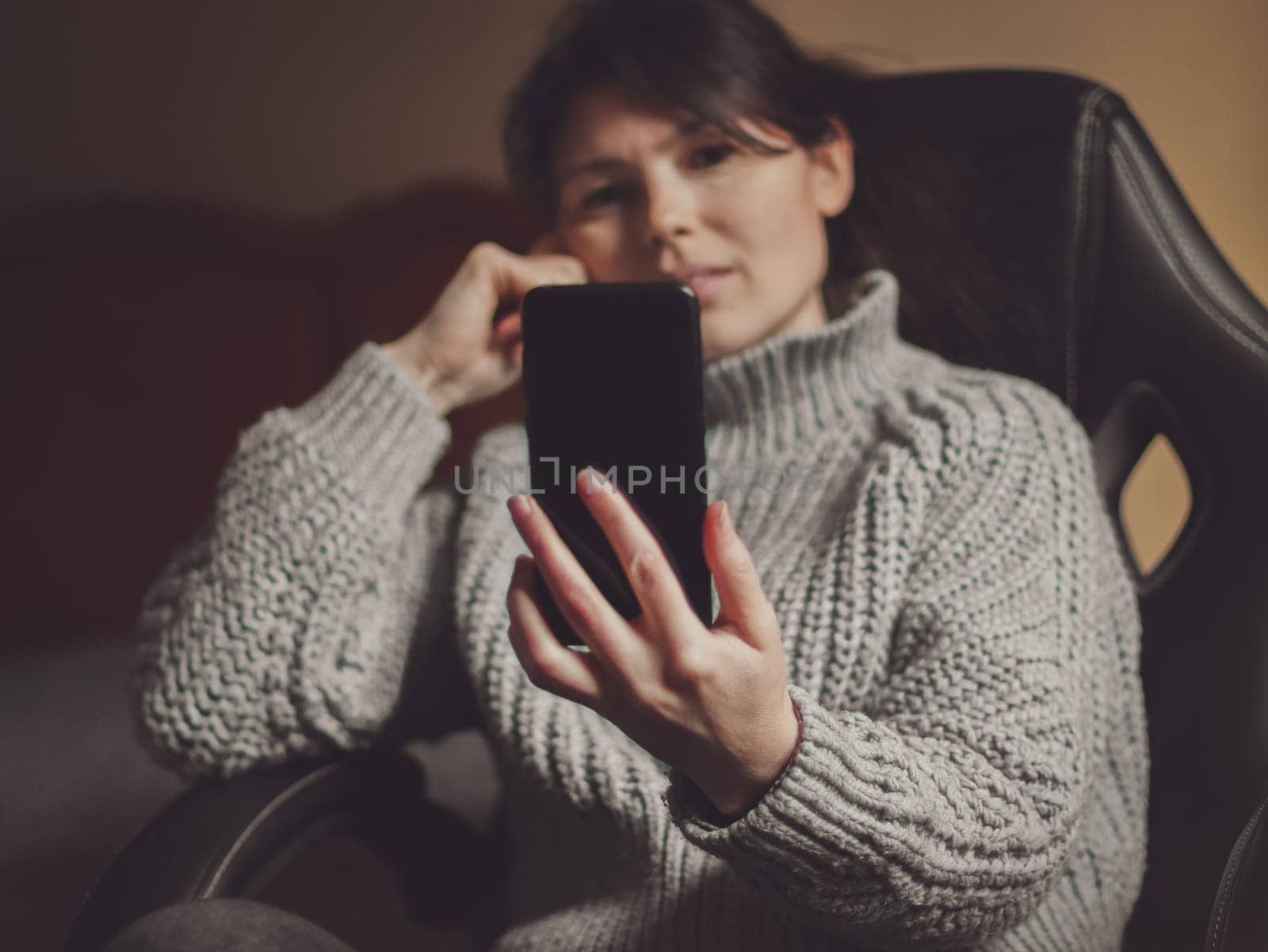 A caucasian young beautiful girl with brown hair and in a thick gray knitted sweater sits in her bedroom on an office chair and chats online with her friend holding a black smartphone in front of her and smiling supports her face with her hand, side view close-up with depth of field. Online chatting concept , social media, modern lifestyle.