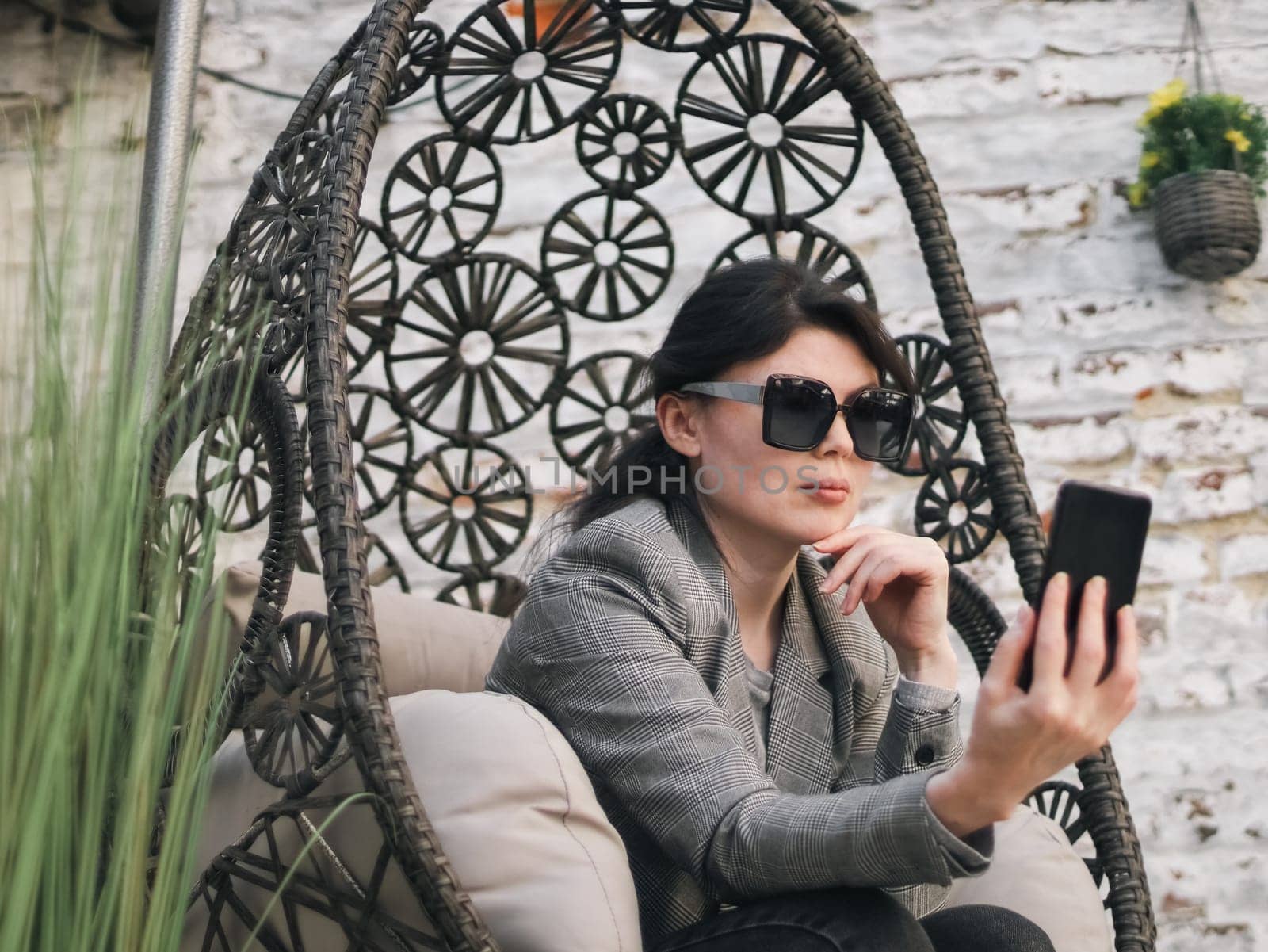 A young beautiful Caucasian girl in a plaid jacket, a gray T-shirt, jeans holds and sunglasses holds a smartphone in her hand and looks at the screen with a serious emotion, holding her chin with her fingers, sitting in a hanging chair against the background of an old brick wall in the backyard of her house, side view close-up. Concept using technology, social media, online chatting, modern lifestyle, at home.
