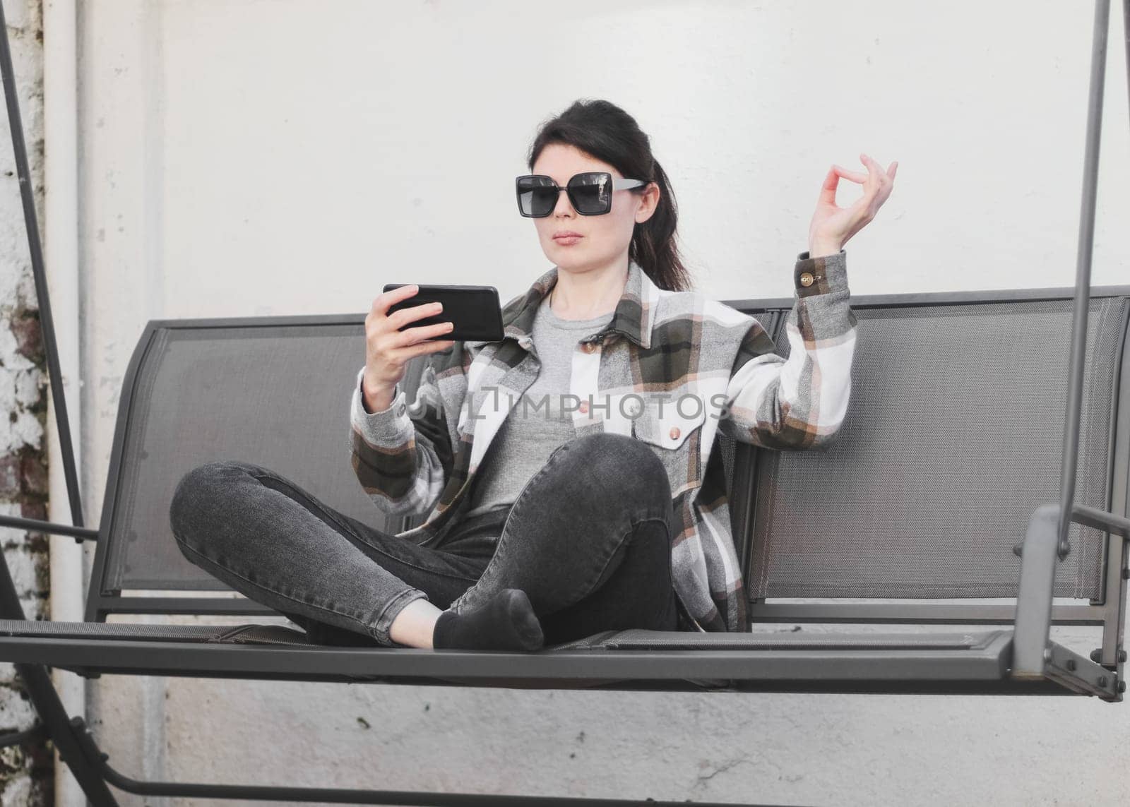 A young beautiful caucasian girl in a plaid shirt, gray jeans and sunglasses holds a smartphone in her hand and learns meditation online, sitting on a garden swing against a white wall in the backyard of her house, close-up side view. Concept using technology, social media, online chats, modern lifestyle, at home, social media, online meditation, course.