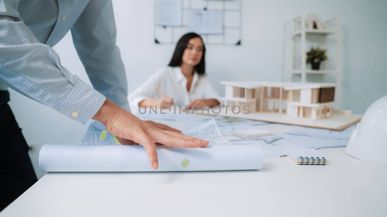 Skilled caucasian interior designer working with house model while discussing with asian architect engineer about house plan on meeting table with blueprint and house model scatter around. Immaculate.