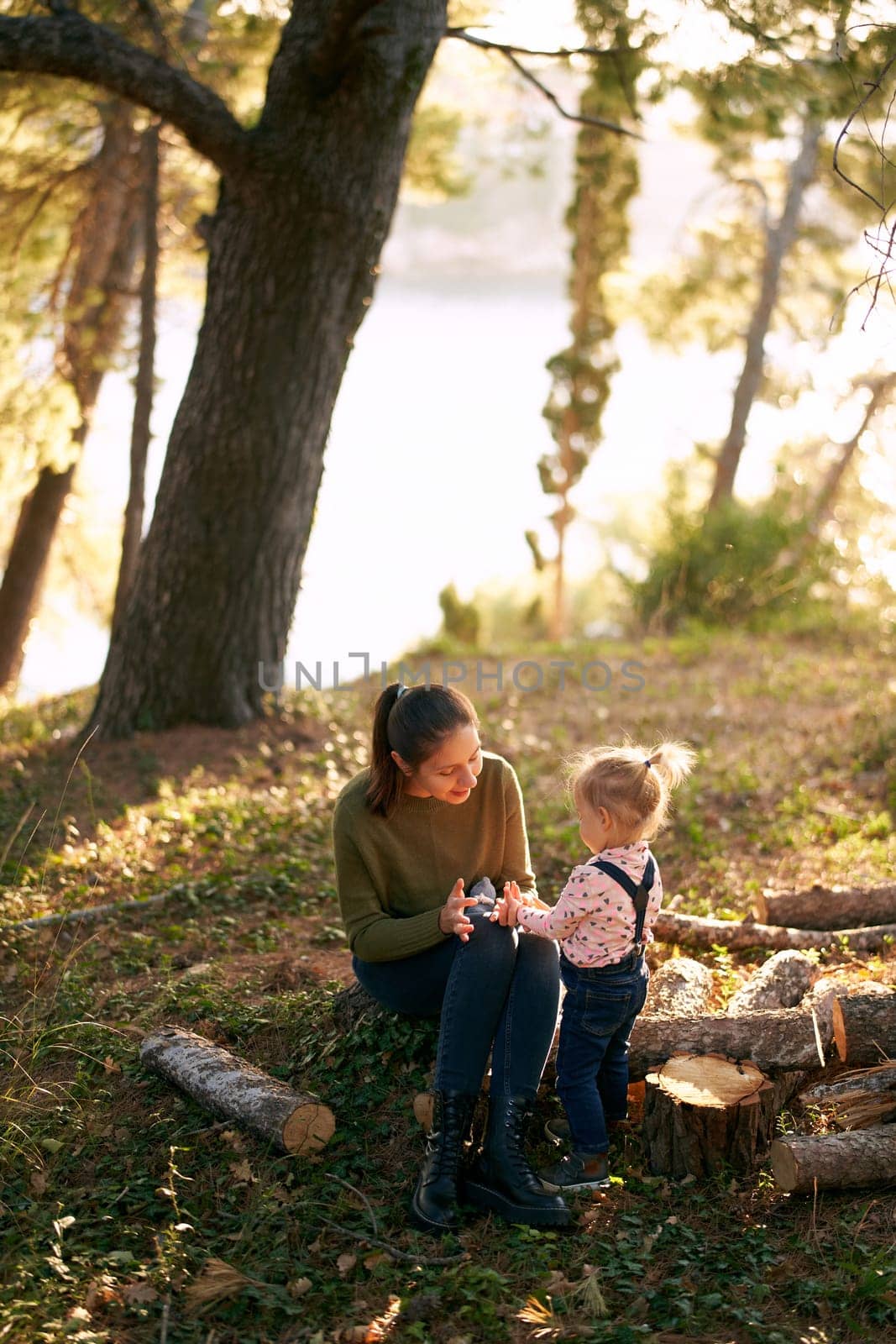 Little girl stands near her mother sitting on a stump in the forest and plays with a toy. High quality photo