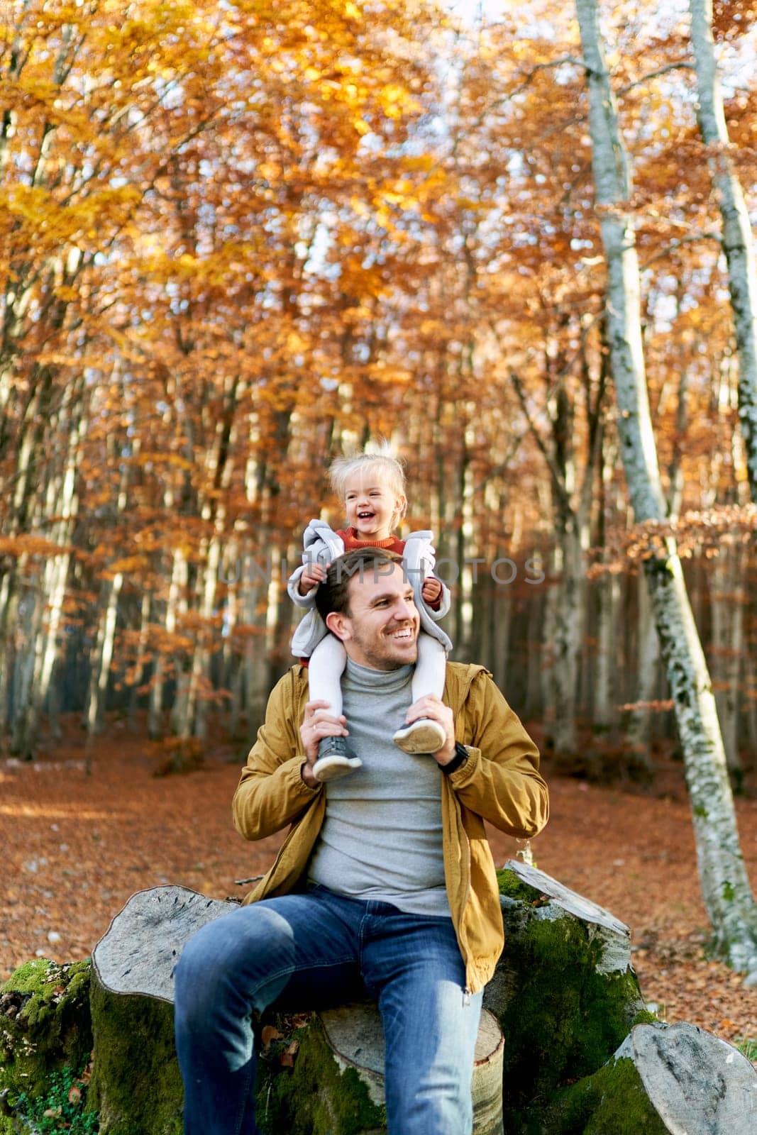Little smiling girl sits on the shoulders of her dad sitting on a stump in the forest. High quality photo