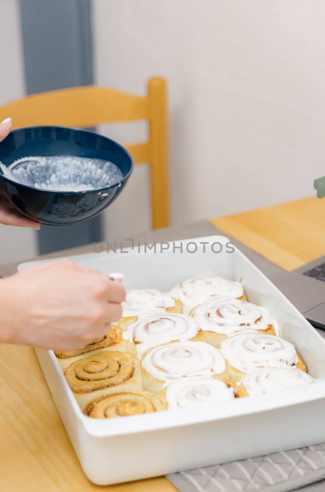 One unrecognizable young Caucasian girl spreads white cream on round hot buns with brown sugar and cinnamon in a white baking dish on a wooden table, standing in the kitchen, close-up side view. Step-by-step instructions for baking synabons. Step 13