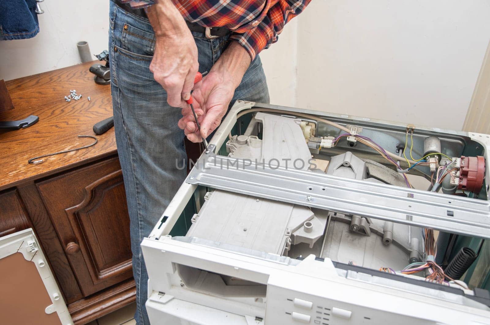 middle-aged master in a checkered suit unscrews the lid of a washing machine with a screwdriver for repair, repairs washing machines and household appliances at home, high quality photo