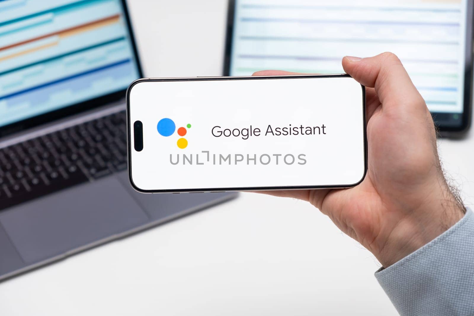 Google Assistant application logo on the screen of smart phone in mans hand, laptop and tablet on the table by vladimka