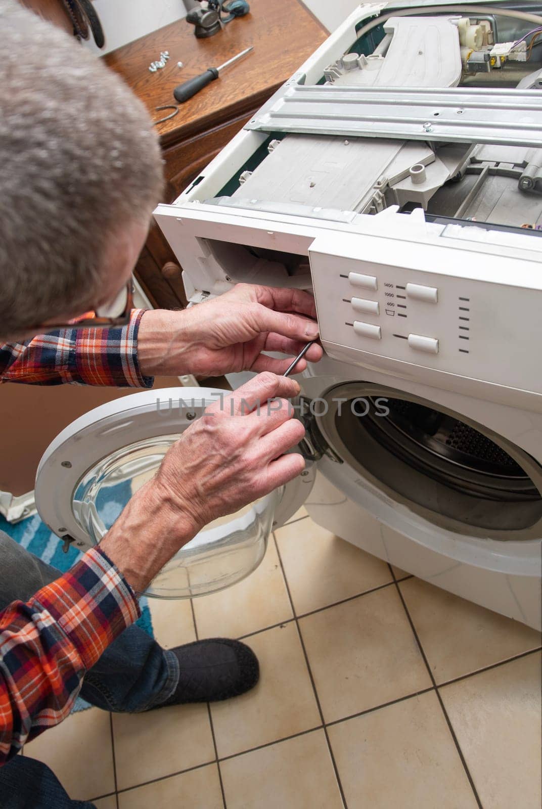 middle-aged master in a checkered suit unscrews the lid of a washing machine with a screwdriver for repair, repairs washing machines and household appliances at home, high quality photo