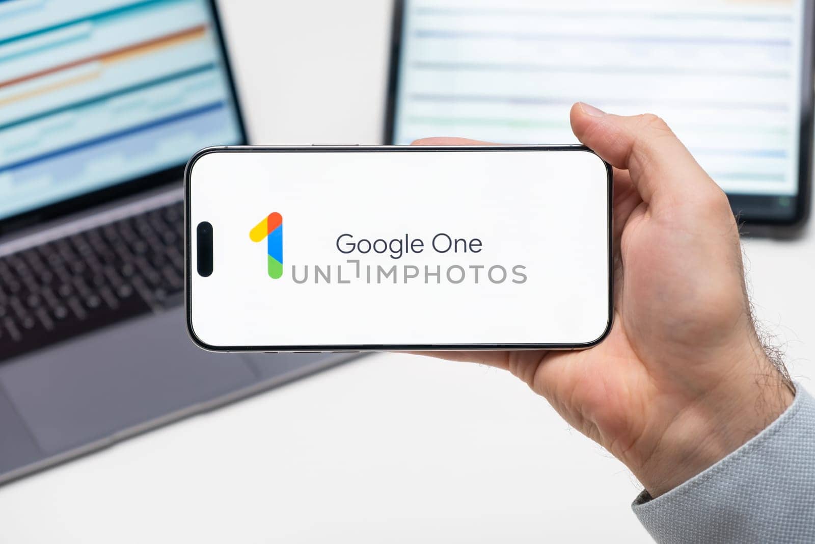 Google One logo of app on the screen of mobile phone held by man in front of the laptop and tablet by vladimka