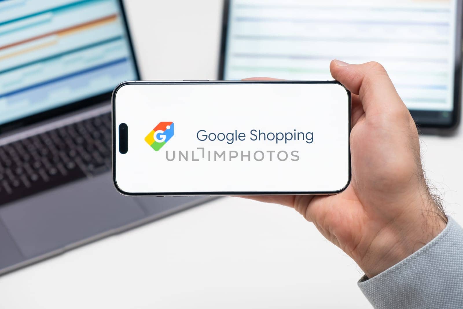 Google Shopping logo of app on the screen of mobile phone held by man in front of the laptop and tablet by vladimka