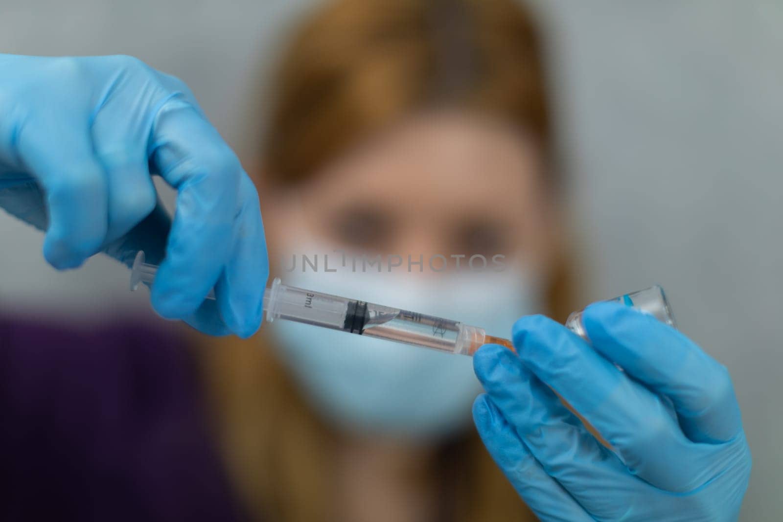 Close-up of the woman's face and hands. She wears disposable gloves and a surgical mask. The woman scoops the preparation into a syringe. Her face is blurred and her hands are sharpened in the foreground.