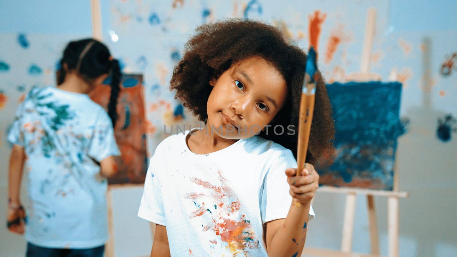 Cute african girl holding painted brush while student drawing canvas behind. Multicultural children attend in art lesson while pretty learner looking at camera. Creative activity concept. Erudition.