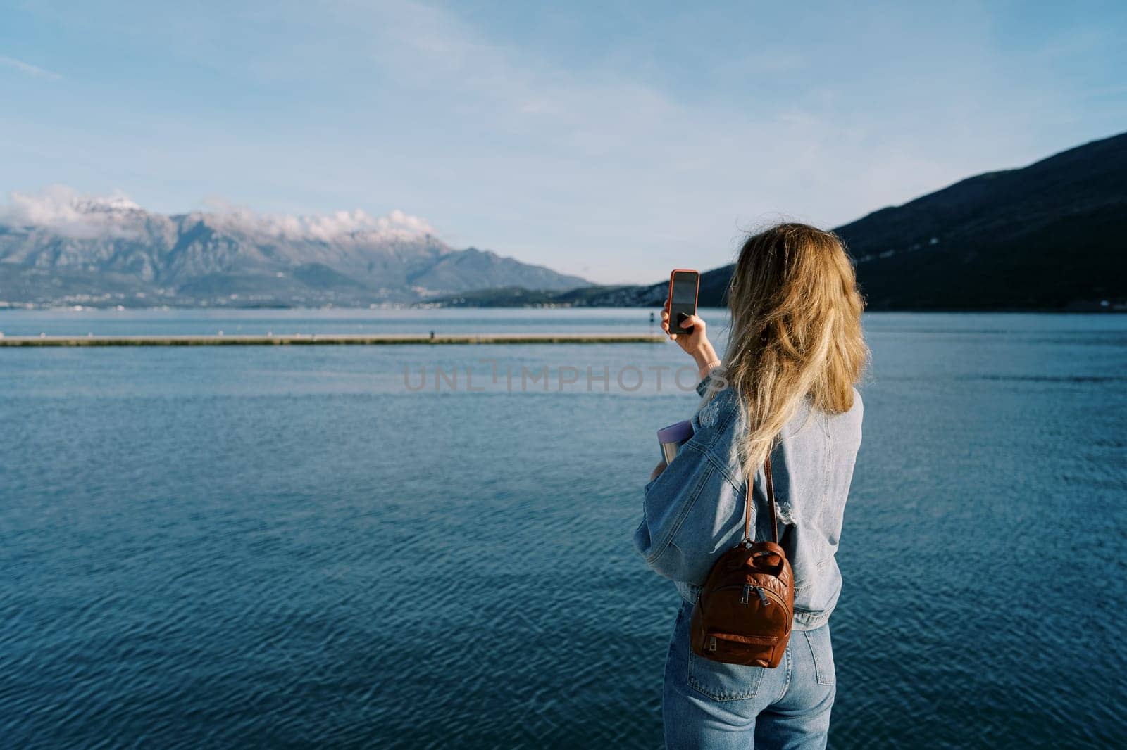 Woman shoots a sea and a mountain range with a smartphone while standing on the shore. Back view by Nadtochiy