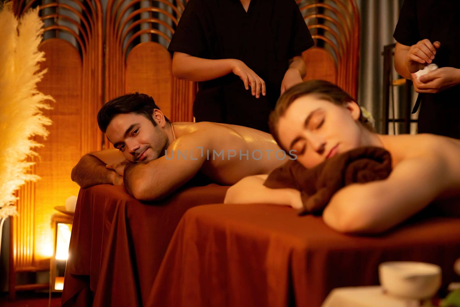 Caucasian couple customer enjoying relaxing anti-stress spa massage and pampering with beauty skin recreation leisure in warm candle lighting ambient salon spa at luxury resort or hotel. Quiescent